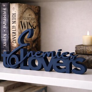 May include: A navy blue wooden sign that reads "Enemies to Lovers" is displayed on a white shelf. The sign is in front of a collection of books, a candle, and a decorative figurine.