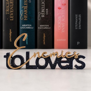May include: A decorative sign with the words "Enemies to Lovers" in gold and navy blue, placed in front of a row of hardcover books. The books have various titles and colors, creating a literary theme.