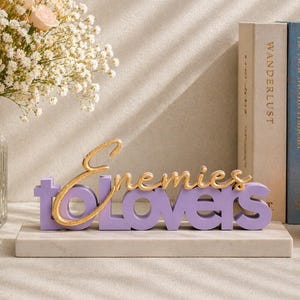 May include: A decorative sign with the words "Enemies to Lovers" in gold and purple. The sign is on a white marble base, with books and a candle in the background. A vase of flowers is on the left.