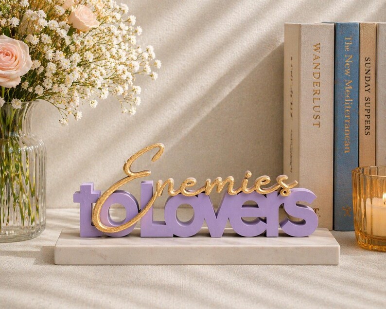May include: A decorative sign with the words "Enemies to Lovers" in gold and purple. The sign is on a white marble base, with books and a candle in the background. A vase of flowers is on the left.