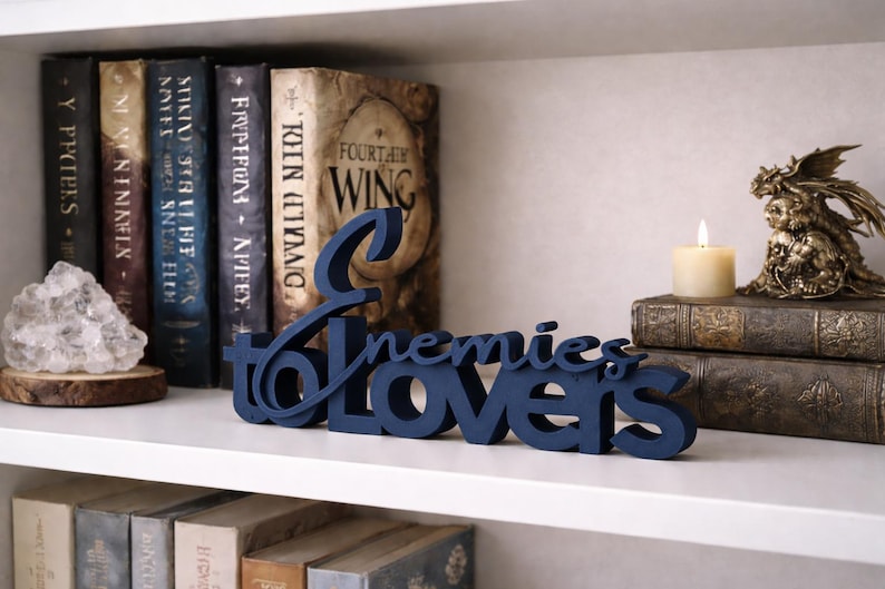 May include: A navy blue wooden sign that reads "Enemies to Lovers" is displayed on a white shelf. The sign is in front of a collection of books, a candle, and a decorative figurine.