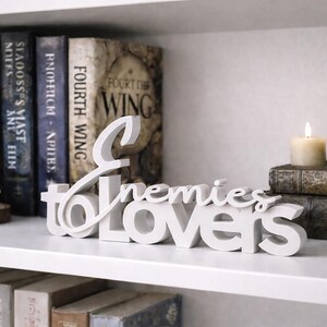 May include: A white wooden sign that reads "Enemies to Lovers" is displayed on a white shelf. The shelf also holds a collection of books, a crystal, a candle, and a decorative figurine.