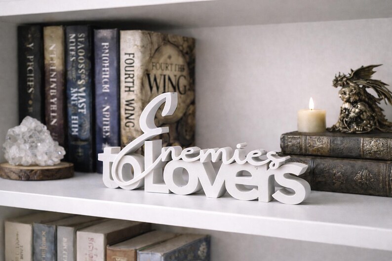 May include: A white wooden sign that reads "Enemies to Lovers" is displayed on a white shelf. The shelf also holds a collection of books, a crystal, a candle, and a decorative figurine.