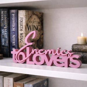 May include: A pink wooden sign that reads "Enemies to Lovers" is displayed on a white shelf. Behind the sign are books, a lit candle, and a decorative figurine. The books have titles like "Fourth Wing".