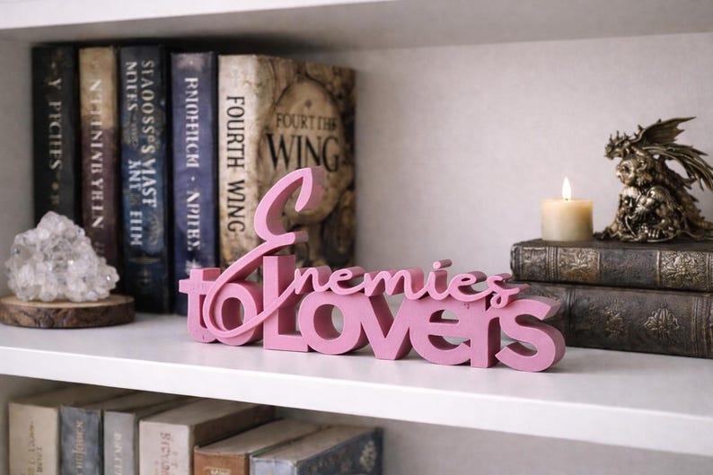 May include: A pink wooden sign that reads "Enemies to Lovers" is displayed on a white shelf. Behind the sign are books, a lit candle, and a decorative figurine. The books have titles like "Fourth Wing".