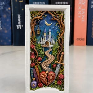 Könnte beinhalten: Ein weißes, rechteckiges Dekorationsobjekt mit einer Fantasy-Szene. Ein Schloss befindet sich unter einem Halbmond und Sternen, umrahmt von Bäumen. Ein Weg führt zum Schloss, mit einem Schwert, einem gebrochenen Herzen und Rosen im Vordergrund.
