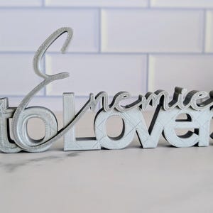 May include: A silver-colored decorative sign that reads "to enemies lovers" in a stylized font. The sign is 3D printed and sits on a light-colored surface, with a white brick background.