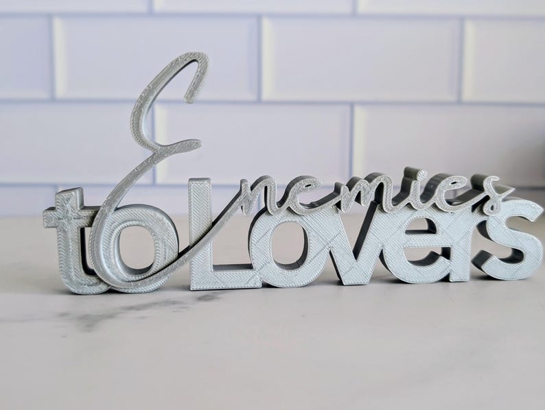 May include: A silver-colored decorative sign that reads "to enemies lovers" in a stylized font. The sign is 3D printed and sits on a light-colored surface, with a white brick background.
