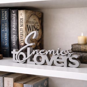 May include: A gray concrete sign that reads "Enemies to Lovers" sits on a white shelf. Behind it are several books with dark spines and a crystal geode on a wooden base. A candle and a dragon statue are also on the shelf.