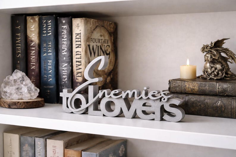 May include: A gray concrete sign that reads "Enemies to Lovers" sits on a white shelf. Behind it are several books with dark spines and a crystal geode on a wooden base. A candle and a dragon statue are also on the shelf.