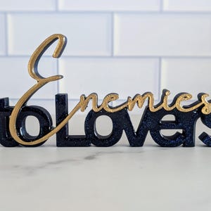 May include: A decorative sign with the words "Enemies to Lovers" in a stylized font. The base of the sign is dark blue with glitter, and the text "Enemies" is in gold. The sign is a decorative item.