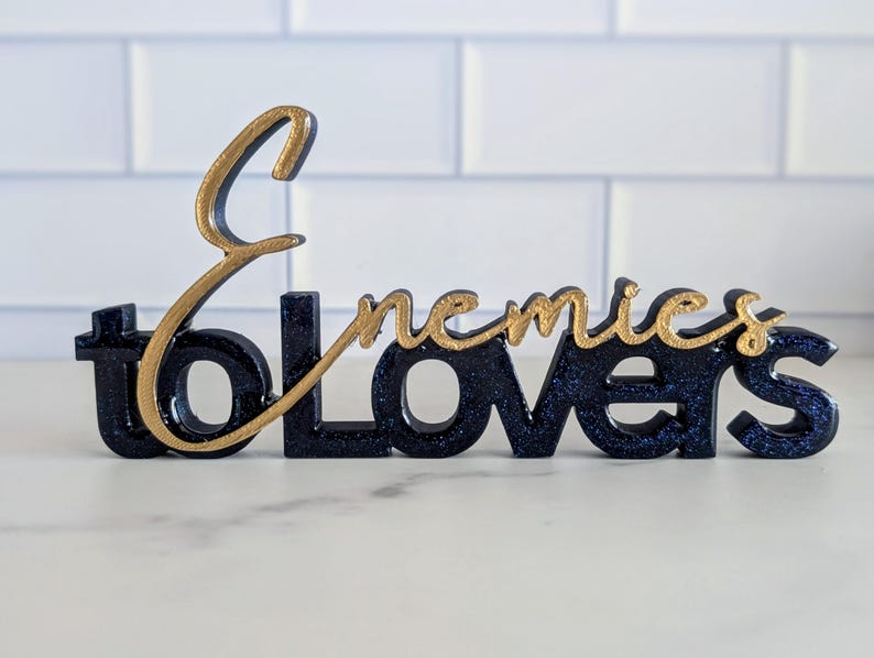 May include: A decorative sign with the words "Enemies to Lovers" in a stylized font. The base of the sign is dark blue with glitter, and the text "Enemies" is in gold. The sign is a decorative item.