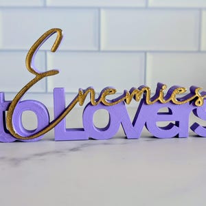 May include: A lavender-colored decorative sign with the words "Enemies to Lovers" in a cursive gold font. The sign is made of a solid material and sits on a white surface.