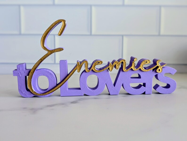 May include: A lavender-colored decorative sign with the words "Enemies to Lovers" in a cursive gold font. The sign is made of a solid material and sits on a white surface.