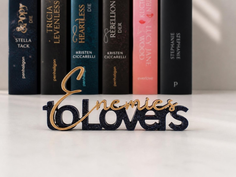 May include: A decorative sign with the words "Enemies to Lovers" in gold and navy blue, placed in front of a row of hardcover books. The books have various titles and colors, creating a literary theme.