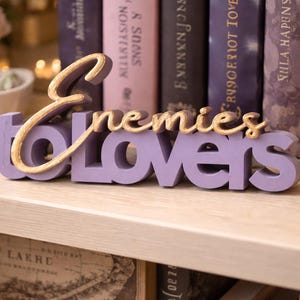 May include: A lavender-colored wooden sign with the words "Enemies to Lovers" in a cursive gold font. The sign is displayed on a light-colored shelf, with books and decorative items in the background.