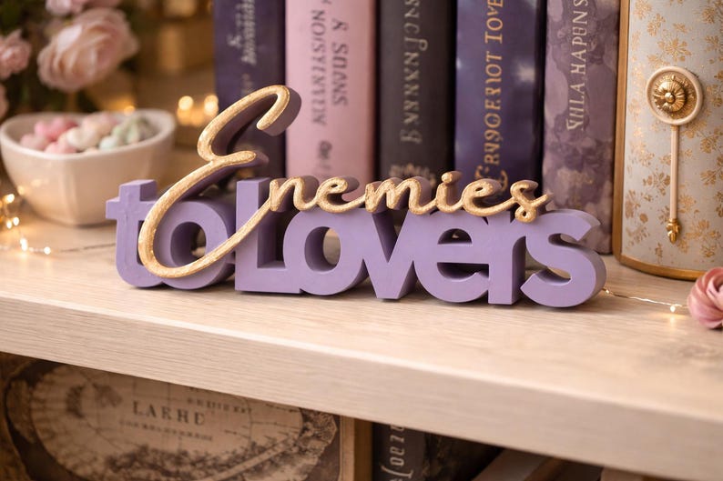 May include: A lavender-colored wooden sign with the words "Enemies to Lovers" in a cursive gold font. The sign is displayed on a light-colored shelf, with books and decorative items in the background.