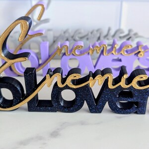 May include: Close-up of three-dimensional word art pieces. The front piece is black with gold script that reads "followers". The second piece is purple with gold script that reads "enemies". The third piece is gray with gold script that reads "enemies".