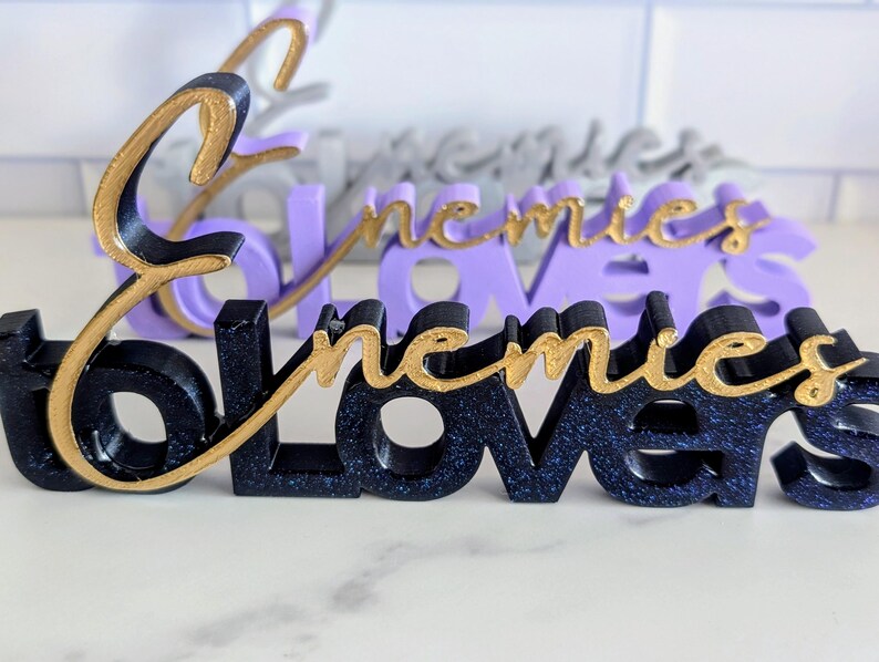 May include: Close-up of three-dimensional word art pieces. The front piece is black with gold script that reads "followers". The second piece is purple with gold script that reads "enemies". The third piece is gray with gold script that reads "enemies".