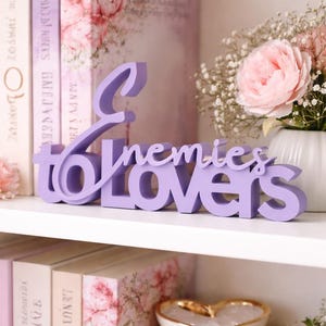 May include: A lavender-colored wooden sign that reads "Enemies to Lovers" is displayed on a white shelf. The sign is in a cursive font. The shelf also holds books, a vase of flowers, and a decorative heart-shaped bowl.