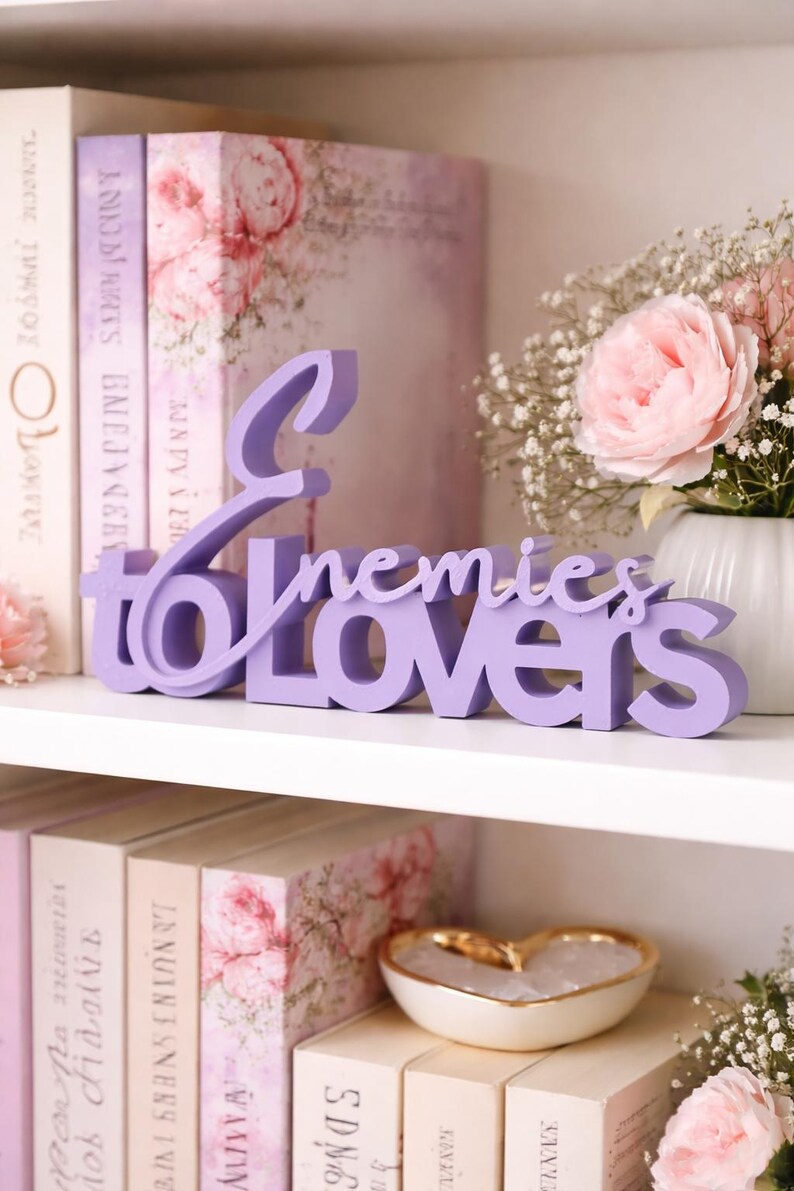 May include: A lavender-colored wooden sign that reads "Enemies to Lovers" is displayed on a white shelf. The sign is in a cursive font. The shelf also holds books, a vase of flowers, and a decorative heart-shaped bowl.