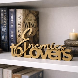 May include: A gold-colored wooden sign that reads "Enemies to Lovers" is displayed on a white shelf. The sign is in front of a collection of books, a lit candle, and a decorative figurine.