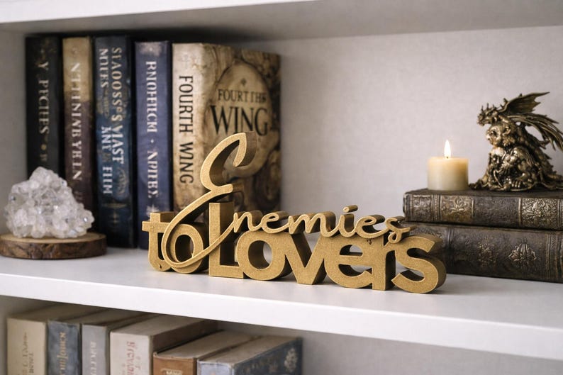 May include: A gold-colored wooden sign that reads "Enemies to Lovers" is displayed on a white shelf. The sign is in front of a collection of books, a lit candle, and a decorative figurine.