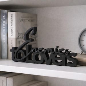May include: A black wooden sign that reads "Enemies to Lovers" in a cursive font. The sign is displayed on a white shelf with books, a gray clock, and a small potted plant. The clock has a white face and black hands.