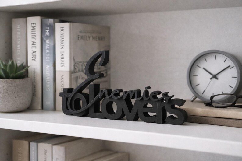 May include: A black wooden sign that reads "Enemies to Lovers" in a cursive font. The sign is displayed on a white shelf with books, a gray clock, and a small potted plant. The clock has a white face and black hands.