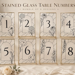 May include: A set of eight rectangular table numbers in gold frames, each featuring a unique black line art floral design and an arched window motif. The text STAINED GLASS TABLE NUMBERS BUNDLE OF 8 DESIGNS is printed above the collection of signs.