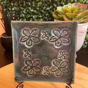 May include: Square ceramic tile with a floral design in shades of green and brown. The tile is displayed on a black metal stand, showcasing the embossed flower pattern. The background features greenery and a wooden surface.