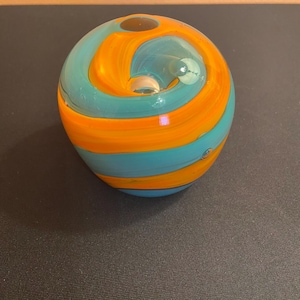 May include: A round, hand-blown glass paperweight with a swirl design. The paperweight features bands of turquoise and orange glass. The glass has a glossy finish and a small opening at the top.