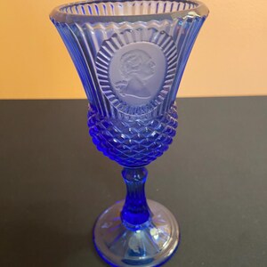 May include: A cobalt blue glass goblet with a detailed design. The goblet features a portrait cameo and diamond-shaped textures. The base is round and the stem is slender. The glass has a fluted rim.