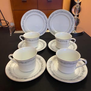 May include: A set of white porcelain teacups and saucers with a delicate gray floral pattern and gold trim. The set includes four cups and saucers, along with two matching dinner plates. The tableware is arranged on a black surface.