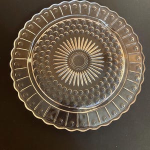 May include: Clear glass serving plate with a scalloped edge. The plate features a central sunburst design surrounded by small, raised dots and radial lines. The plate is round.