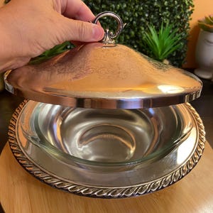 May include: A silver-toned metal serving dish with a removable glass bowl insert. The metal lid features an ornate engraved scroll pattern and a circular handle. The base of the metal dish has a decorative twisted rope edge design.