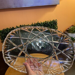 May include: An oval clear glass serving platter featuring a geometric starburst and diamond-cut pattern across its surface. The dish has a smooth, flat center surrounded by a textured, faceted rim that catches light, creating a reflective appearance.