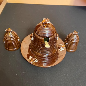 May include: A brown ceramic beehive-shaped honey pot set with two matching salt and pepper shakers. The honey pot has a small opening and a yellow honey dipper. The set is decorated with small bee accents.