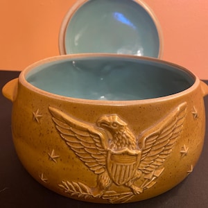 May include: A tan ceramic bowl with a lid, featuring an embossed eagle design and star accents. The interior is a light blue color. The bowl has two handles and is likely for serving.