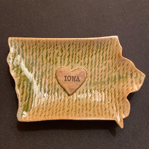 May include: A handmade ceramic dish shaped like the state of Iowa. The dish is glazed in shades of green and brown, with a heart-shaped accent in the center that reads "IOWA". The dish has a textured surface.