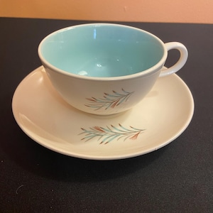 May include: A vintage teacup and saucer set. The cup has a light blue interior and a white exterior with a handle. The saucer and cup are decorated with a sprig design in shades of teal and rust. The set is on a black surface.