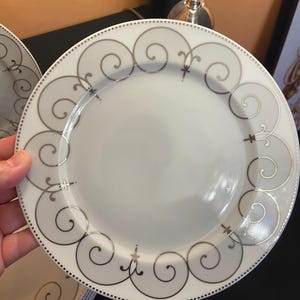 May include: White ceramic plate with a silver scroll design around the rim. The plate has a dotted border and a simple, elegant aesthetic. The plate is part of a dinnerware set.