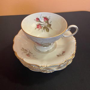 May include: A vintage teacup and saucer set. The cup and saucers are cream-colored with a gold trim and a floral design featuring red roses. The set includes a stack of saucers.