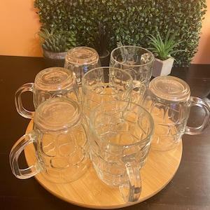 May include: A set of seven clear glass beer mugs with handles, arranged on a round wooden tray. The mugs have a textured, grid-like pattern. The background includes greenery and a dark surface.