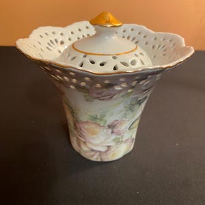 May include: A white porcelain decorative container with a floral design. The container has a flared, openwork rim and a lid with a gold finial. The container is decorated with pink and yellow roses.