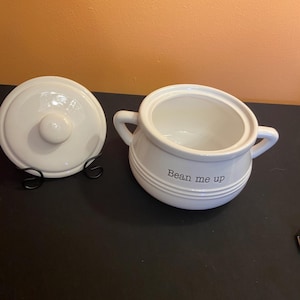 May include: White ceramic soup tureen with lid and handles. The tureen has the phrase "Bean me up" printed on the side. The lid is round with a small knob. The tureen is on a black surface.
