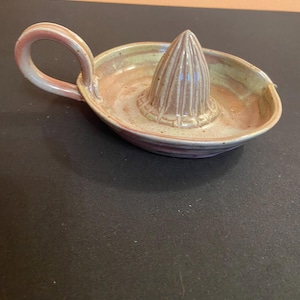 May include: Handmade ceramic citrus juicer in shades of pink, tan, and green. The juicer features a conical reamer in the center, surrounded by a shallow bowl with a handle. The pottery has a rustic, artisanal appearance, perfect for kitchen use.