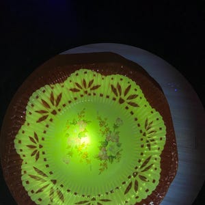 May include: A vintage, round, glowing green glass plate with a brown rim. The plate features a floral design in the center and decorative patterns around the edge. The plate is illuminated, highlighting its details and colors.