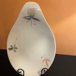 May include: White, teardrop-shaped decorative plate with floral and leaf designs in gray, pink, and yellow. The plate is displayed on a black metal stand against an orange background. The plate has a mid-century modern aesthetic.