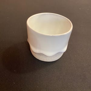 May include: A small, white ceramic cup with a rounded top and a textured, gray base. The white glaze drips down the side, creating a unique, organic shape. The cup is designed for holding liquids.